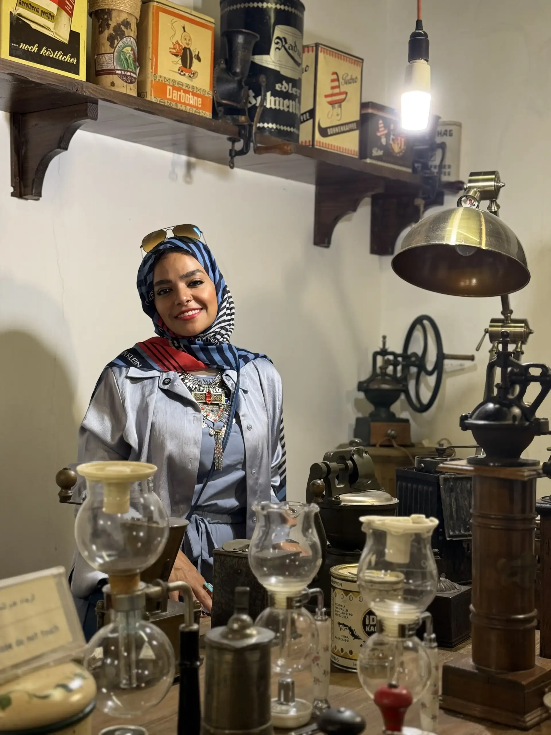 لقطة داخل مقهى تراثي: سيدة تبتسم بين أدوات تحضير قديمة—قصة القهوة و الصحة العامة من مصدرها حتى فنجانك.