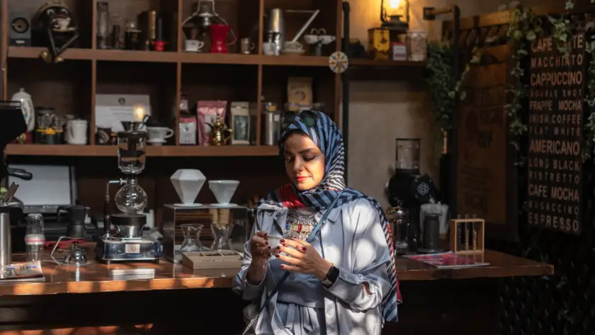 سيدة تستمتع بفنجان القهوة العلاجية في مقهى مختص بإضاءة دافئة وأدوات تحضير يدوية—لحظة هدوء تعزز المزاج والتركيز.