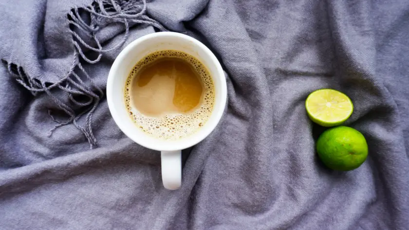 كوب قهوة على قماش رمادي بجانب ليمون أخضر مقطوع—مشهد يعبّر عن القهوة العلاجية بالليمون في المنزل.