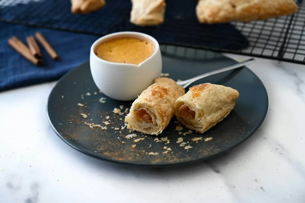 فنجان قهوة كريمية مع لفائف مخبوزة على طبق—تقديم صباحي أنيق مستوحى من ترند القهوة بزيت الزيتون.