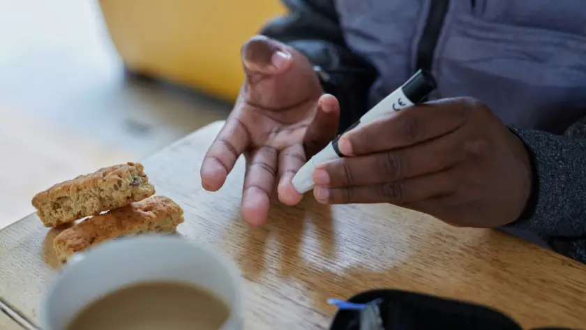 شخص يقيس سكر الدم بجهاز فحص بجانب كوب قهوة ووجبة خفيفة—توضيح لعلاقة القهوة للصحة العامة وسكر الدم.