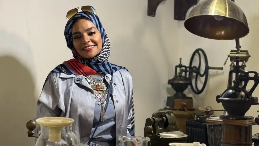 لقطة داخل مقهى تراثي: سيدة تبتسم بين أدوات تحضير قديمة—قصة القهوة و الصحة العامة من مصدرها حتى فنجانك.